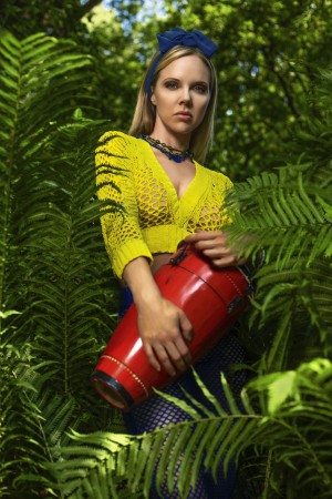 Beautiful young blonde woman wearing a colorful summer outfit holding a red djembe on the edge of a jungle